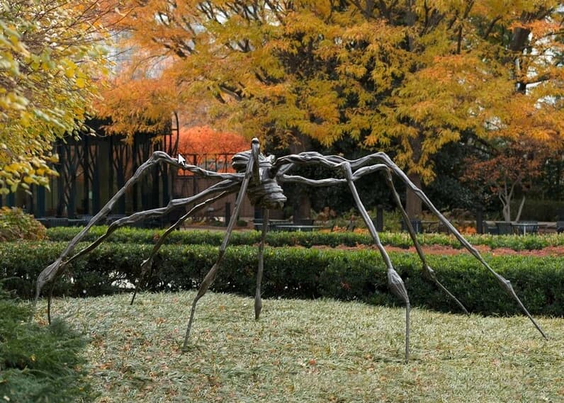 A large bronze spider sculpture in a garden beneath trees with yellow and orange autumn leaves
